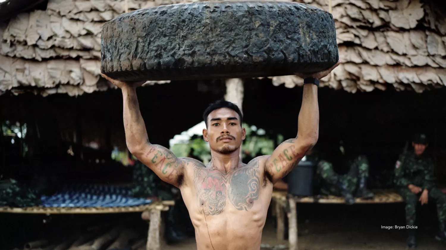 A tattooed Burmese man holds a boulder over his head while staring stoically at the viewer. The tattoos on both his upper arms read "Peace"