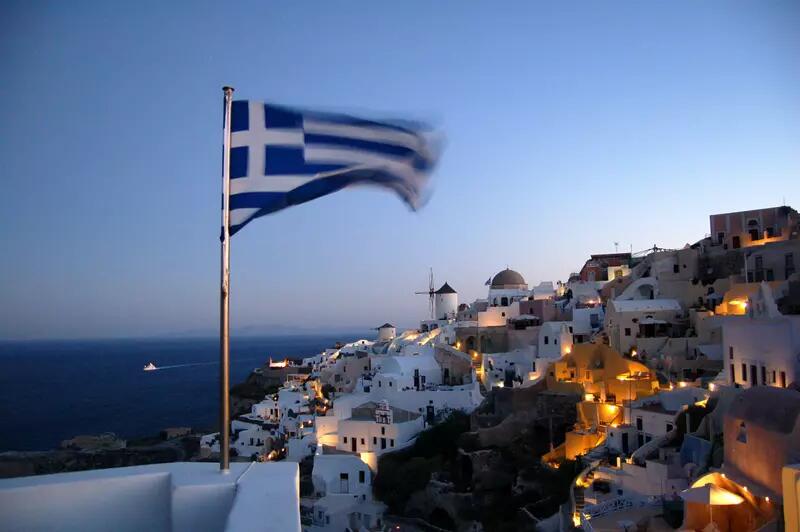 A Greek flag attached to a wall, with a hillside village and the sea in the background