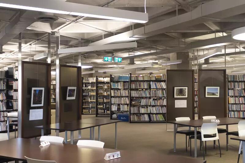 A view of the library, with tables and chairs, metal screens displaying framed photos, and behind them rows of bookshelves.