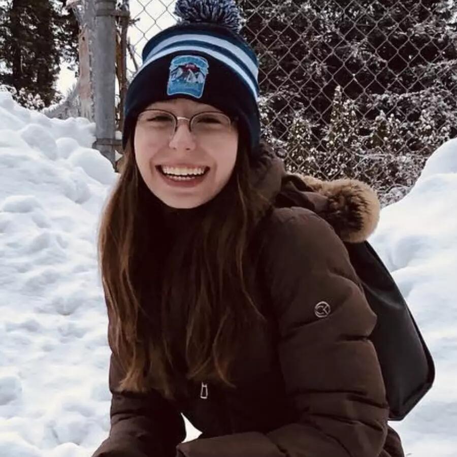Woman wearing glasses and a beanie crouching down in the snow and smiling.