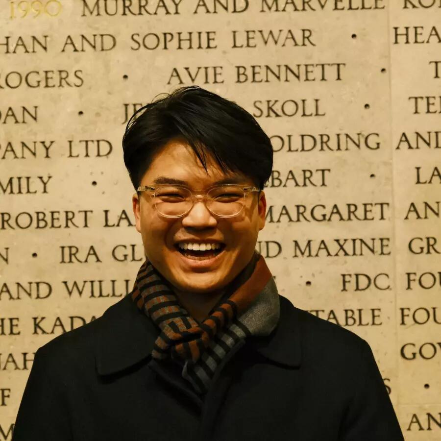 Man wearing glasses, a scarf, and winter coat, standing in front of a wall inscribed with names. 